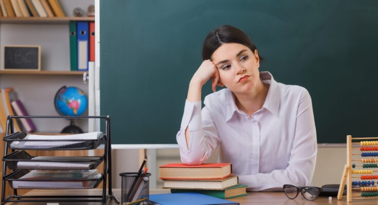 stockking-freepik-cansada-poner-mano-mejilla-joven-maestra-sentada-escritorio-herramientas-escolares-aula-scaled